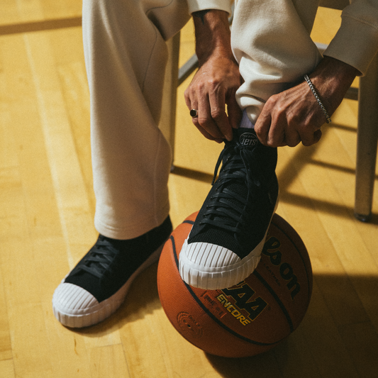A person wearing black high-top Red Ball Jets sneakers and cream sweatpants sits on a gym floor, resting one foot on a basketball.
