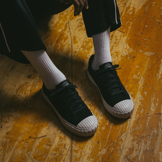 A person wearing black low-top Red Ball Jets sneakers with white crew socks sits on a worn wooden gym floor, feet side by side.