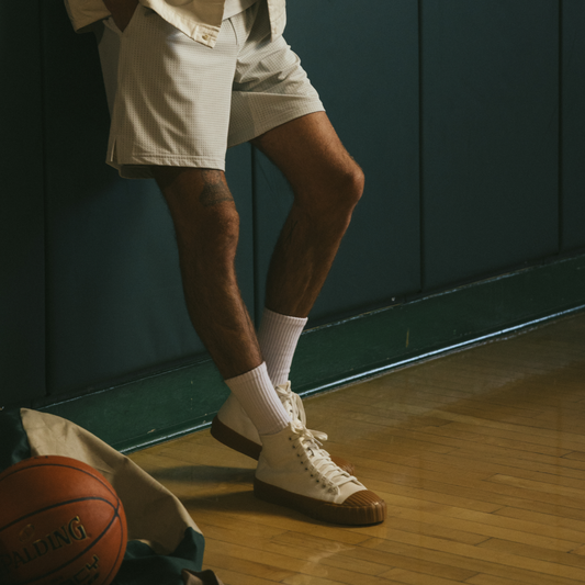 A person wearing off-white high-top Red Ball Jets sneakers with gum soles leans casually against a dark gym wall. They are dressed in a light-colored jacket and shorts, with hands in pockets. A few basketballs and a gym bag rest on the polished wood floor beside them, lit by warm natural light.