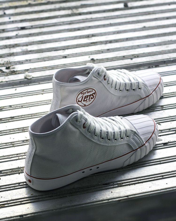 White Legacy HI sneakers resting on metal bleacher steps, showing the heel patch with the Red Ball Jets logo and the ribbed rubber sole detail along the side.