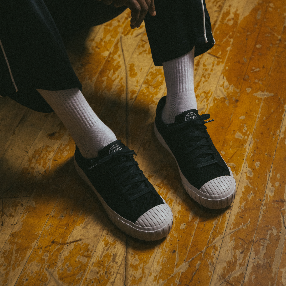 A person wearing black low-top Red Ball Jets sneakers with white crew socks sits on a worn wooden gym floor, feet side by side.
