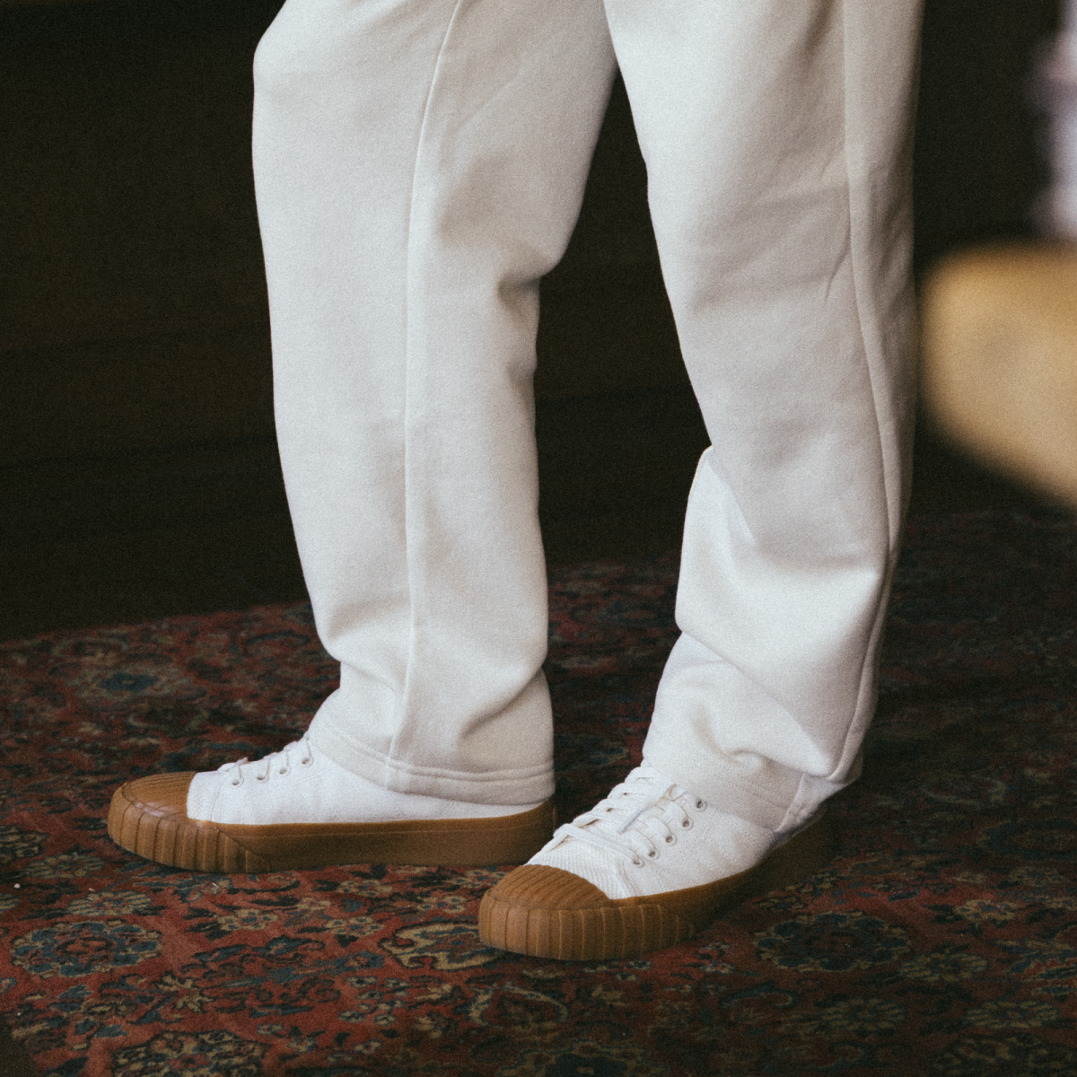 A person wearing off-white low-top Red Ball Jets sneakers with gum soles and white sweatpants stands on a vintage patterned rug.
