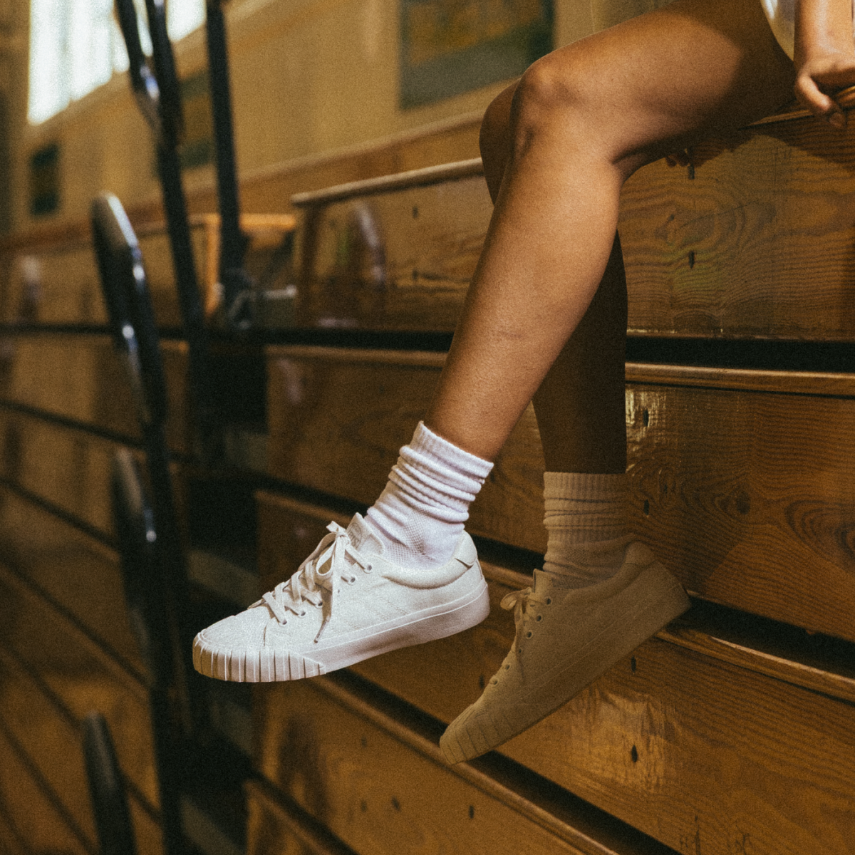 A person wearing white low-top Red Ball Jets sneakers and white crew socks sits on wooden bleachers, legs relaxed in natural light.