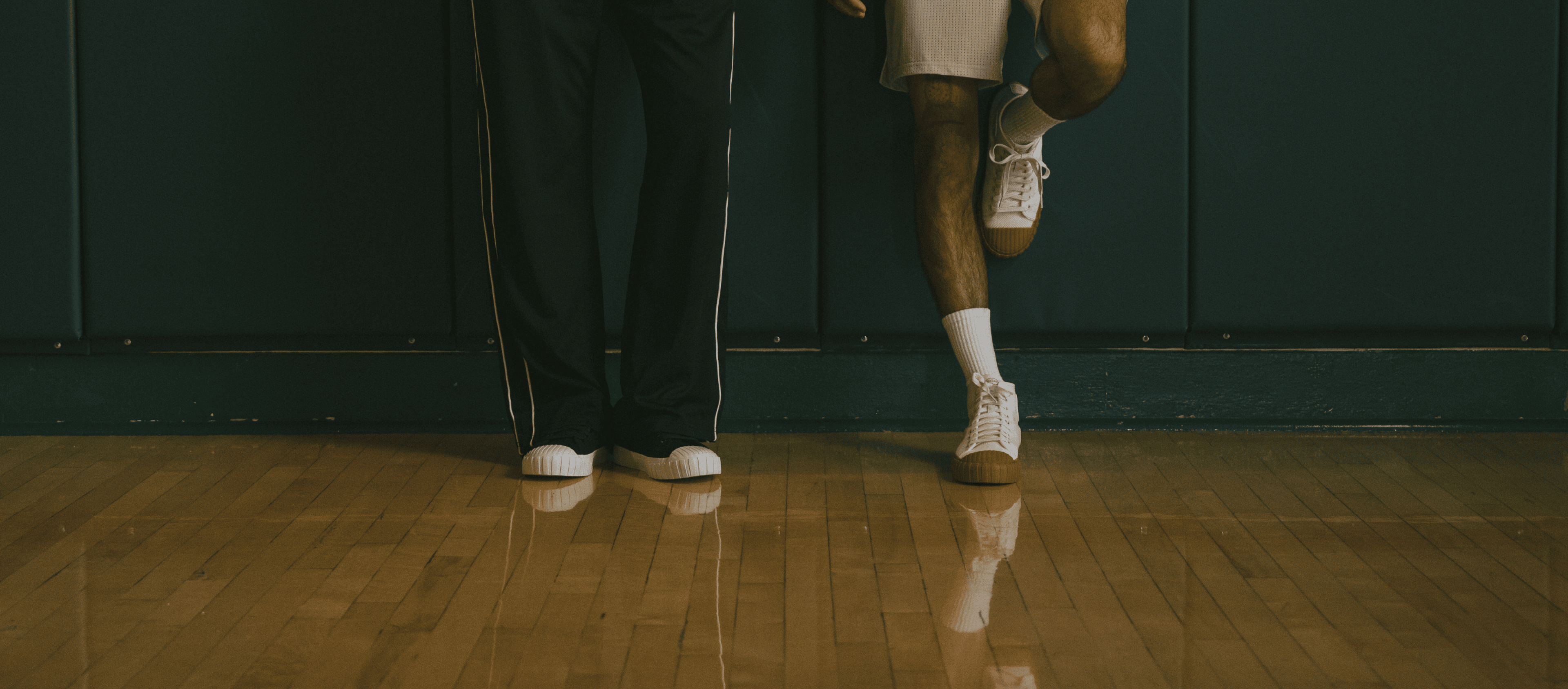 Two people standing on a wooden floor with a dark wall in the background, with one person's leg lifted.