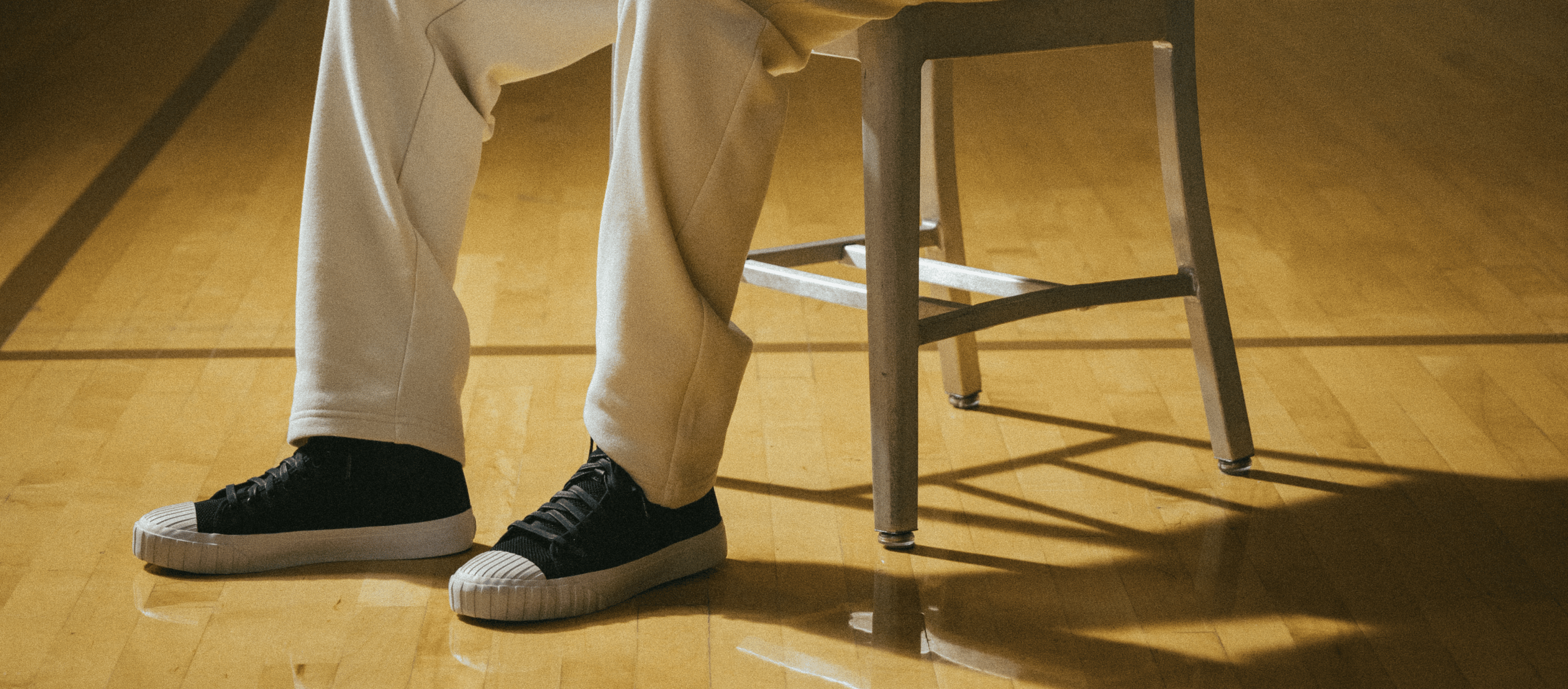 Person wearing black sneakers and white pants sitting on a chair with wooden floor.