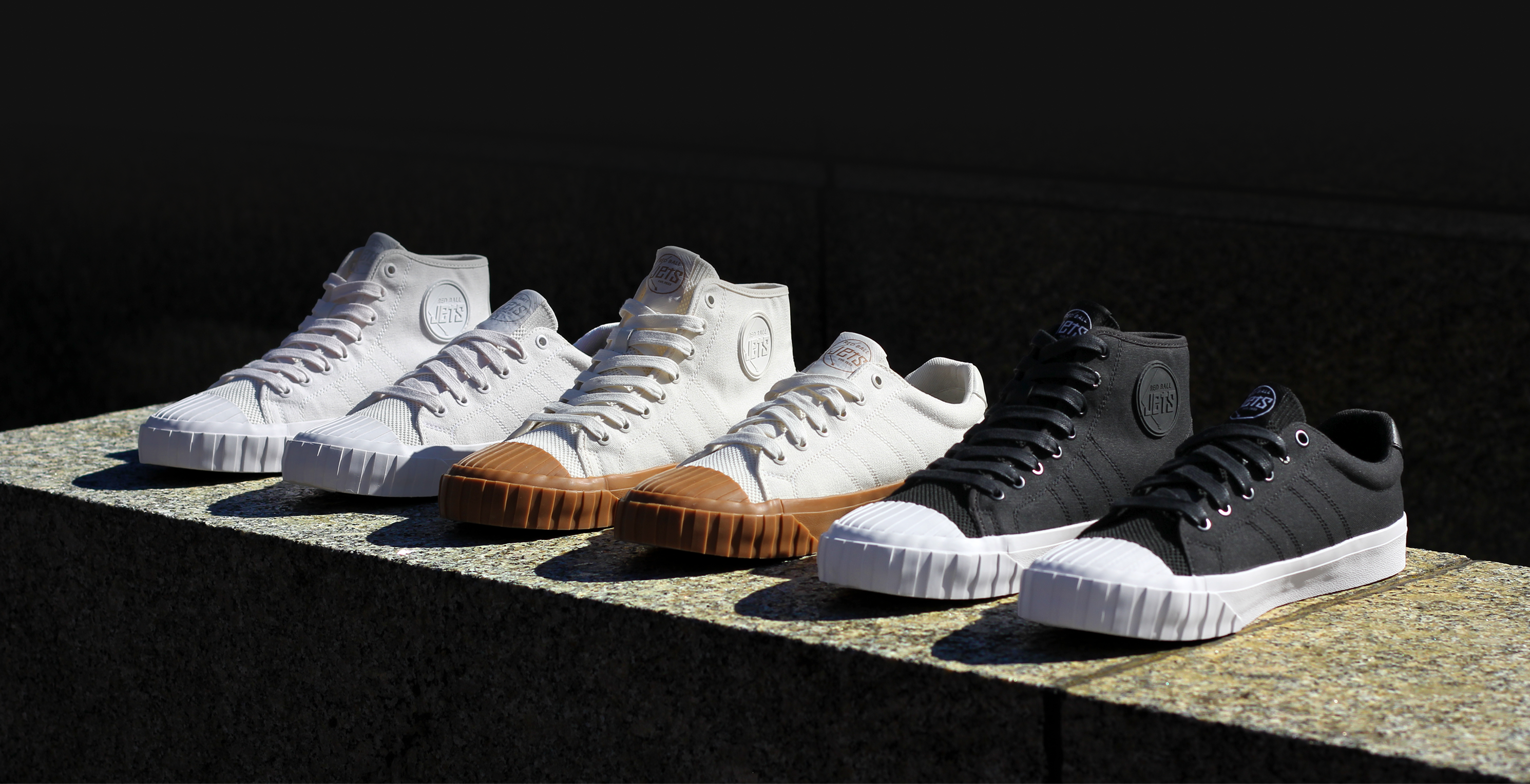 Four pairs of sneakers in white, brown, and black on a stone surface with a dark background.