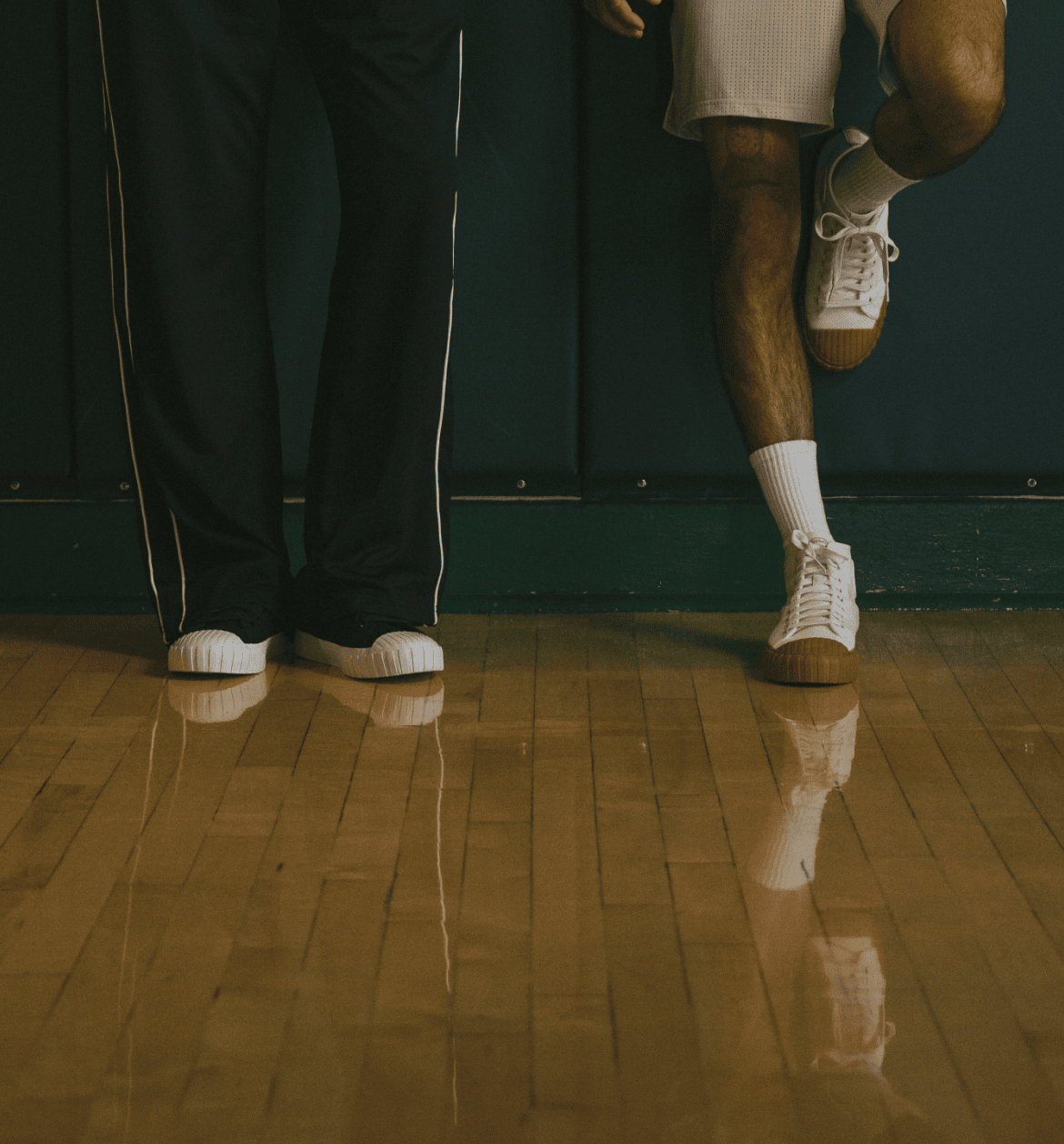 Two people standing on a wooden floor with one person lifting a leg.