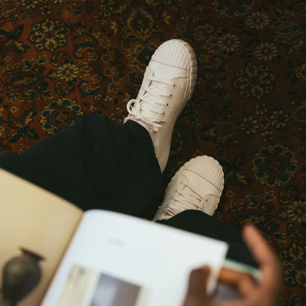 All white Red Ball Jets' low-top sneakers on the subjects feet, below the book they're reading in a book store.