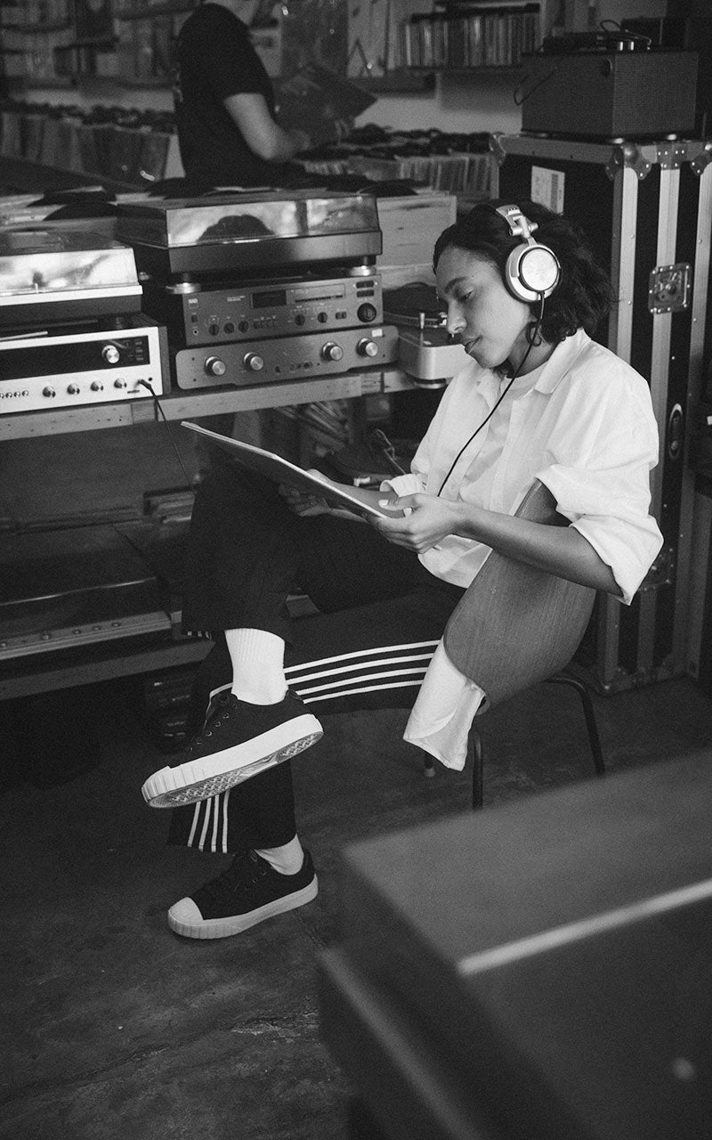Black and white photo of a young black woman sitting in a record shop listening-station, looking an an LP cover, while liking right in her Red Ball Jet's black low-tops